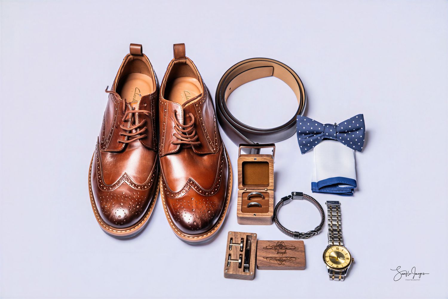 Brown leather brogue shoes with matching belt, blue bow tie, gold watch, wallet, and accessories flat lay on white background.