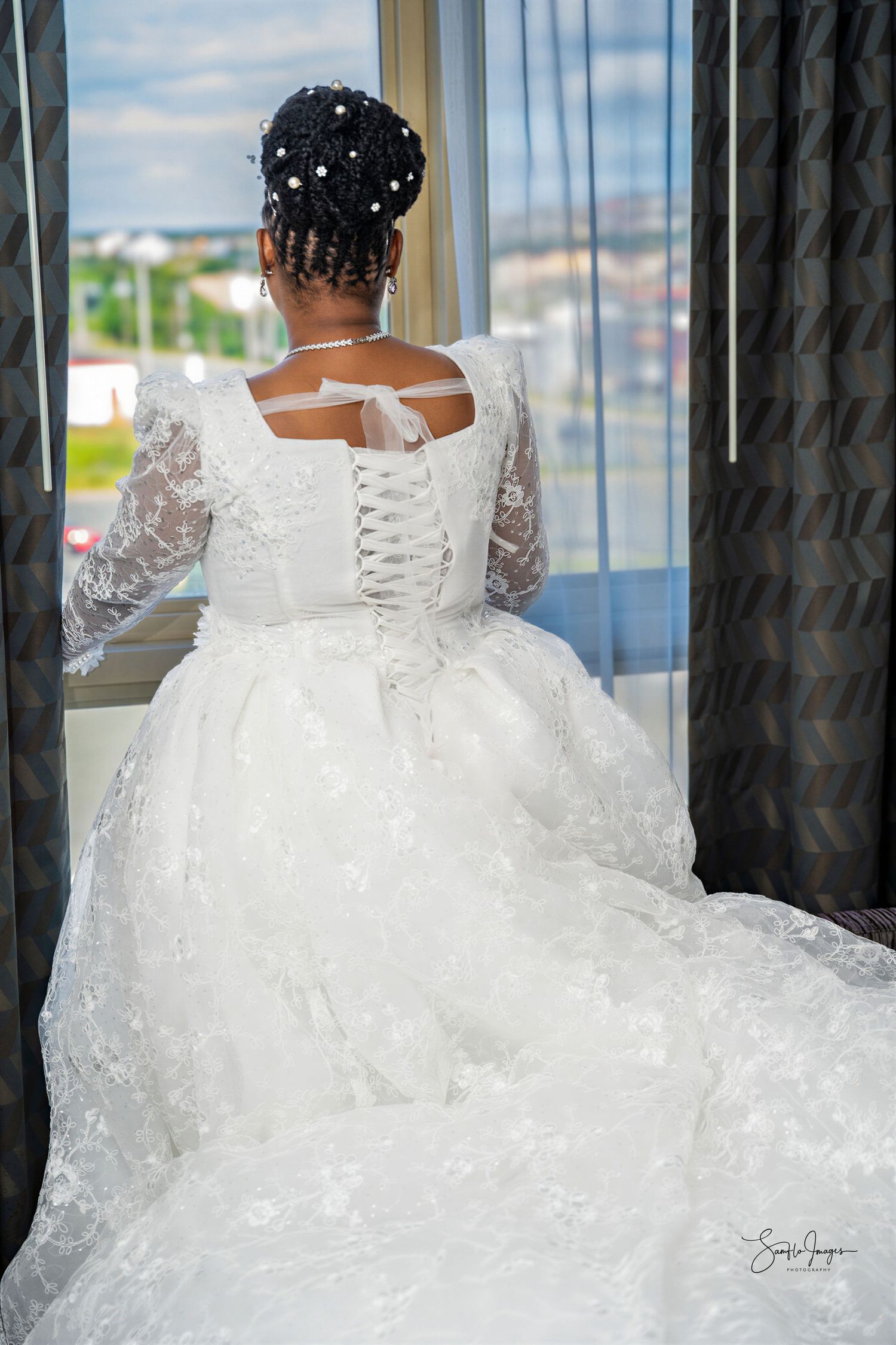 Bride in white lace wedding gown with corset back and long train stands by window, hair adorned with pearl pins.