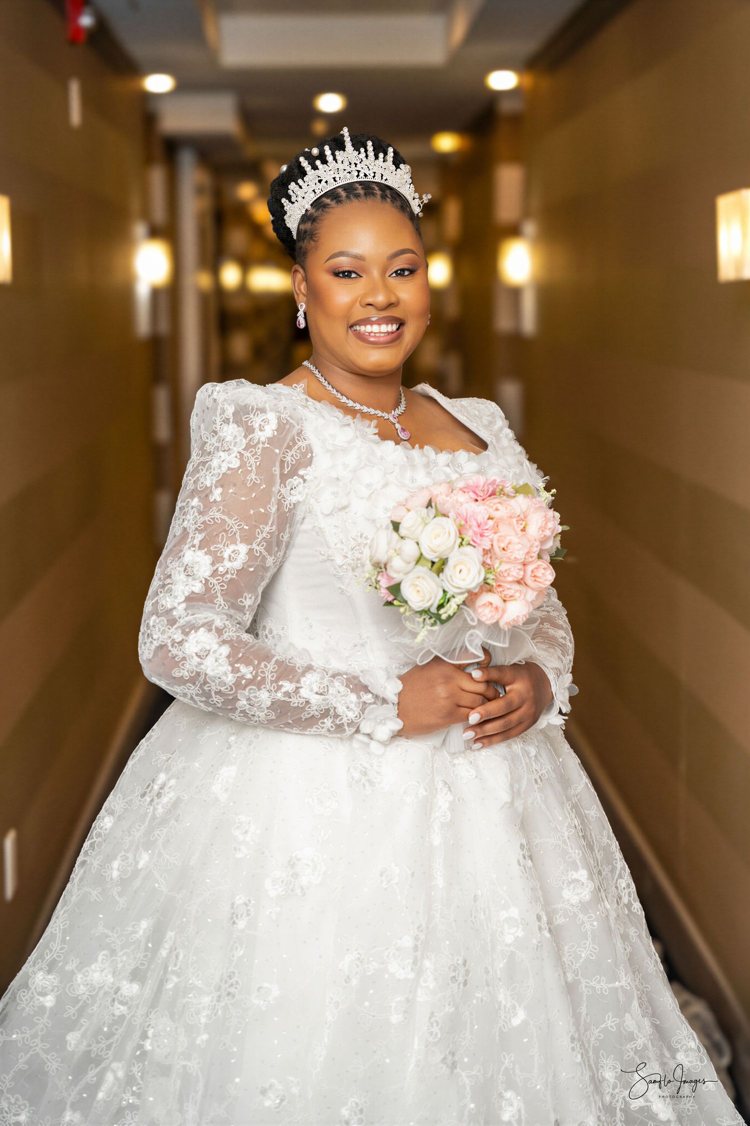 Smiling bride in white lace ball gown with long sleeves, crystal tiara, pearl necklace, holding pink and white bouquet in hotel hallway.