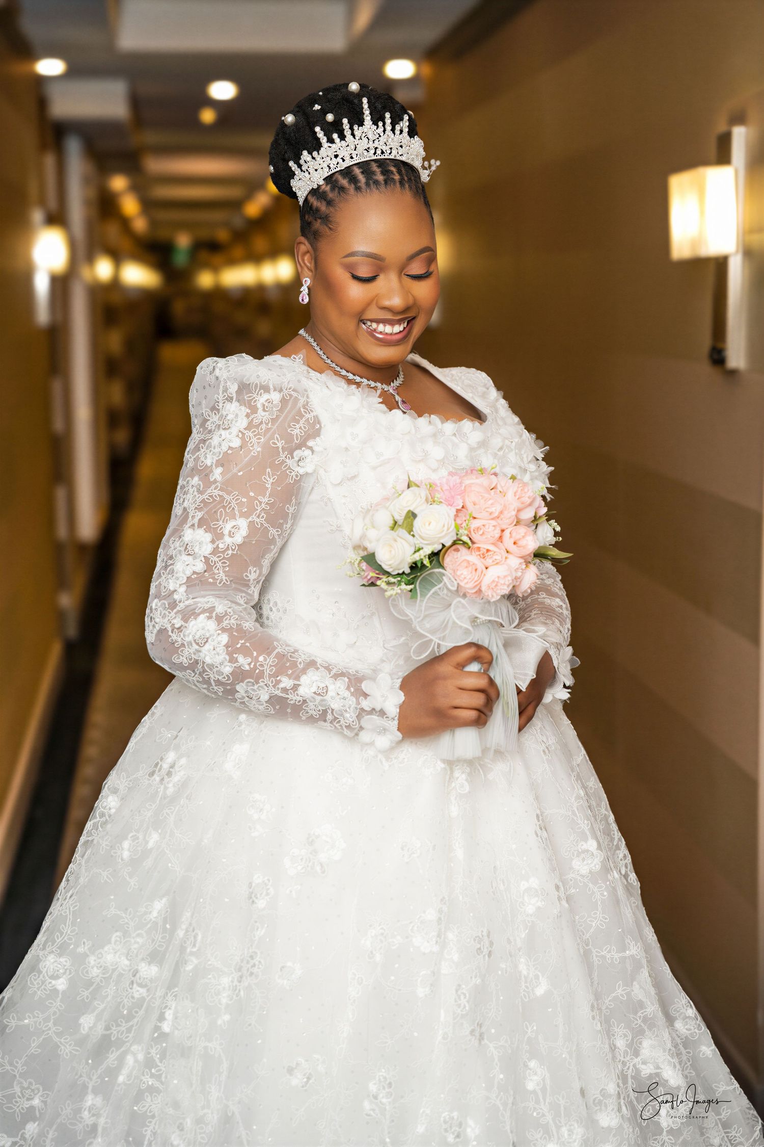 Smiling bride in white lace ball gown with tiara holds pink bouquet in elegant hallway.