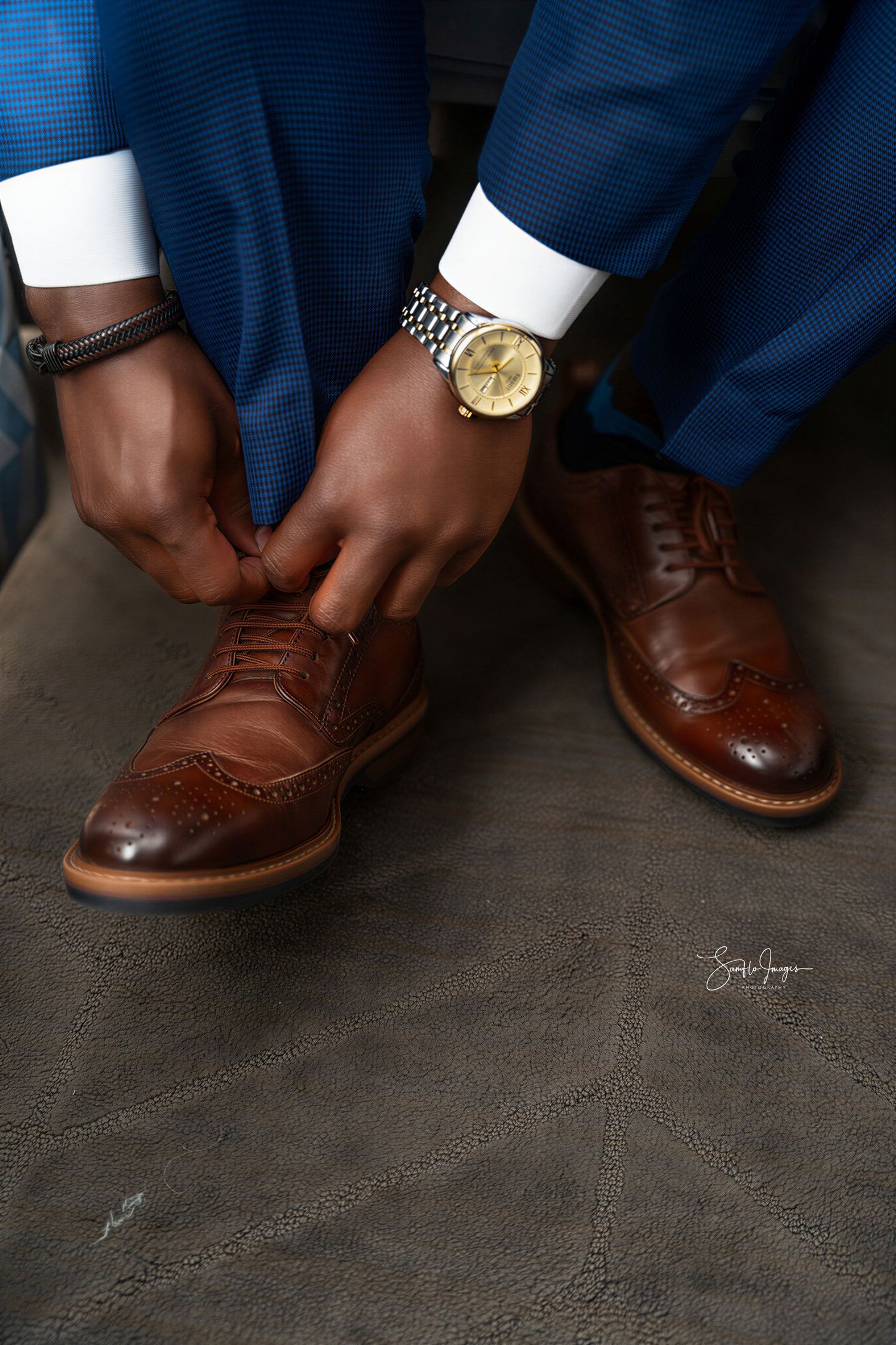 Man in blue suit tying brown wingtip brogue dress shoes while wearing a gold watch and bracelet.