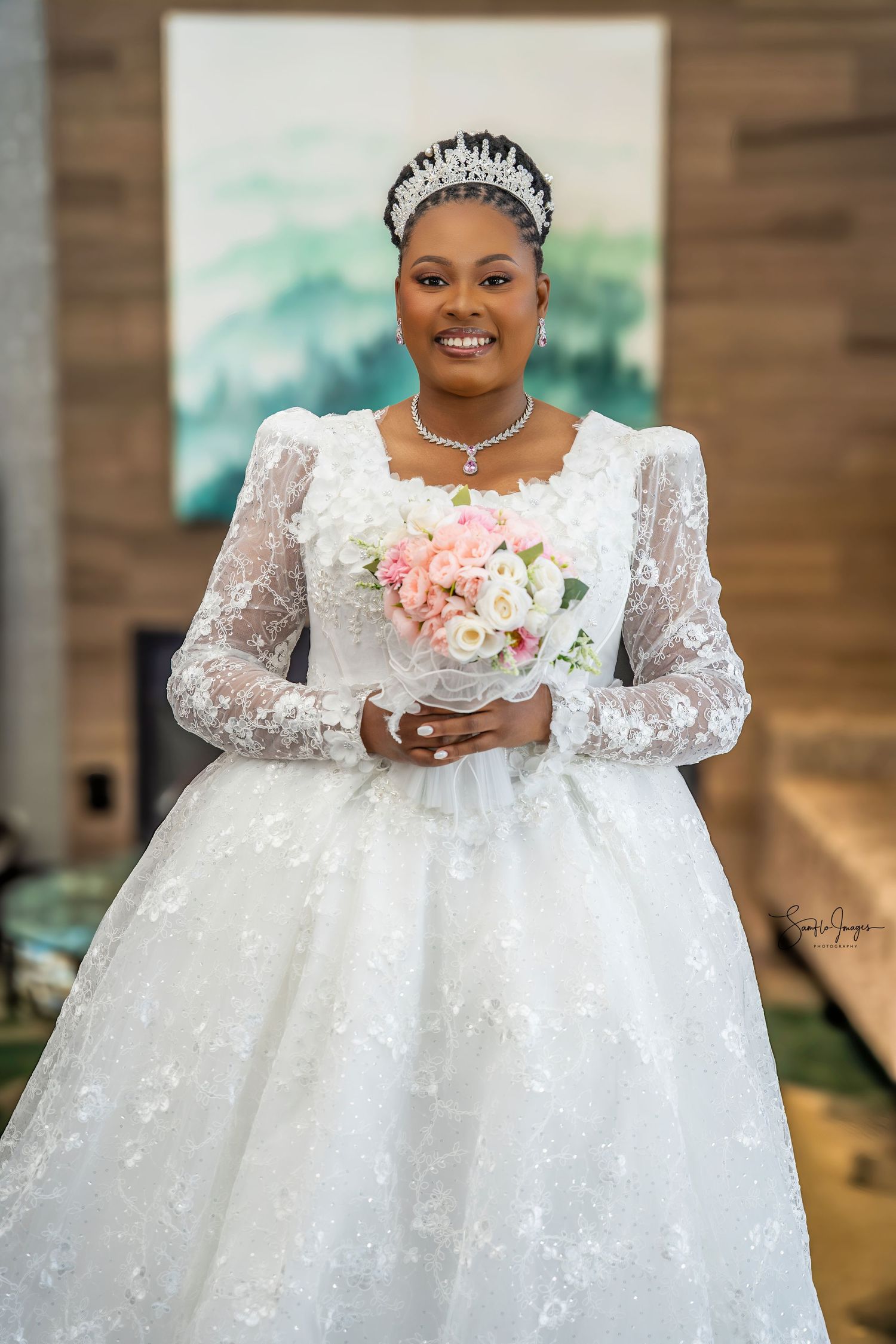 Smiling bride in white lace gown with tiara holds pink bouquet at elegant indoor wedding venue.