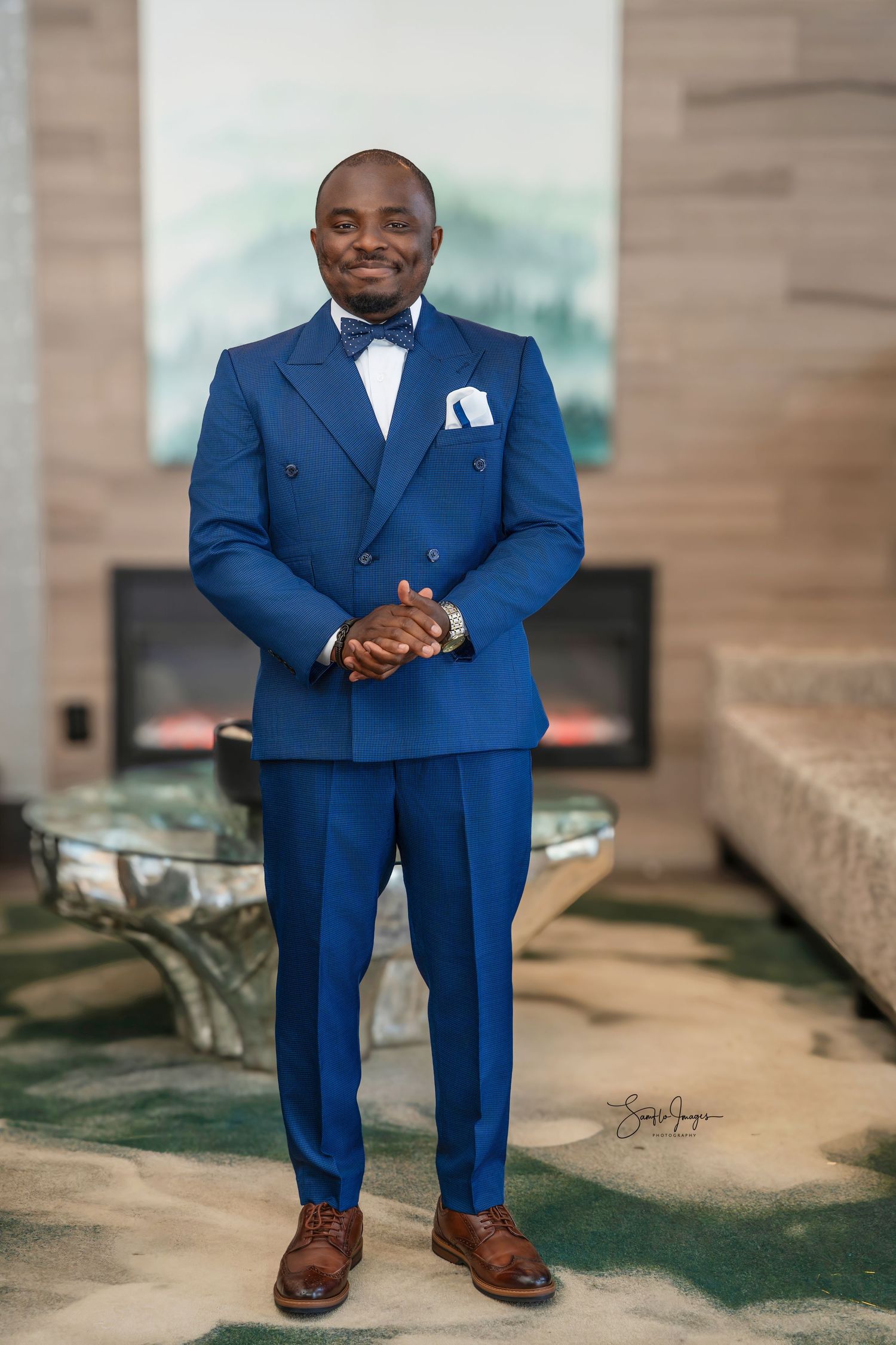 Man in blue double-breasted suit and bow tie smiling indoors near fireplace.