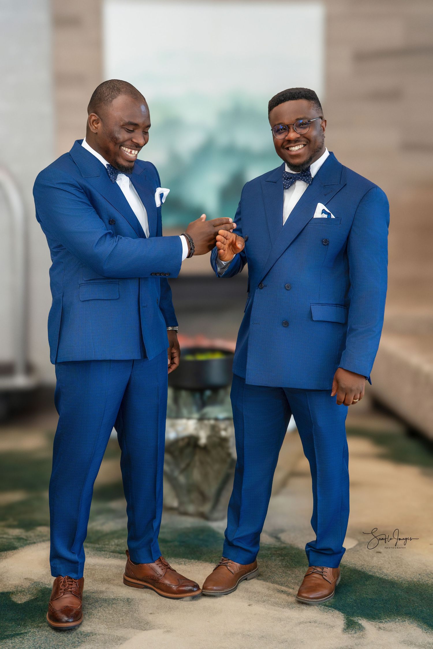 Two men in matching royal blue suits and bow ties share a joyful handshake, smiling warmly at a formal event.
