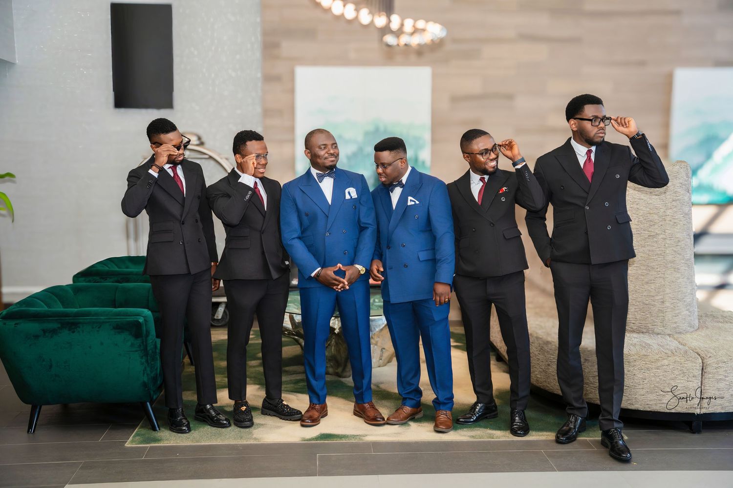 Groom in blue suit poses with five groomsmen in dark suits, raising glasses in a toast at a wedding venue.