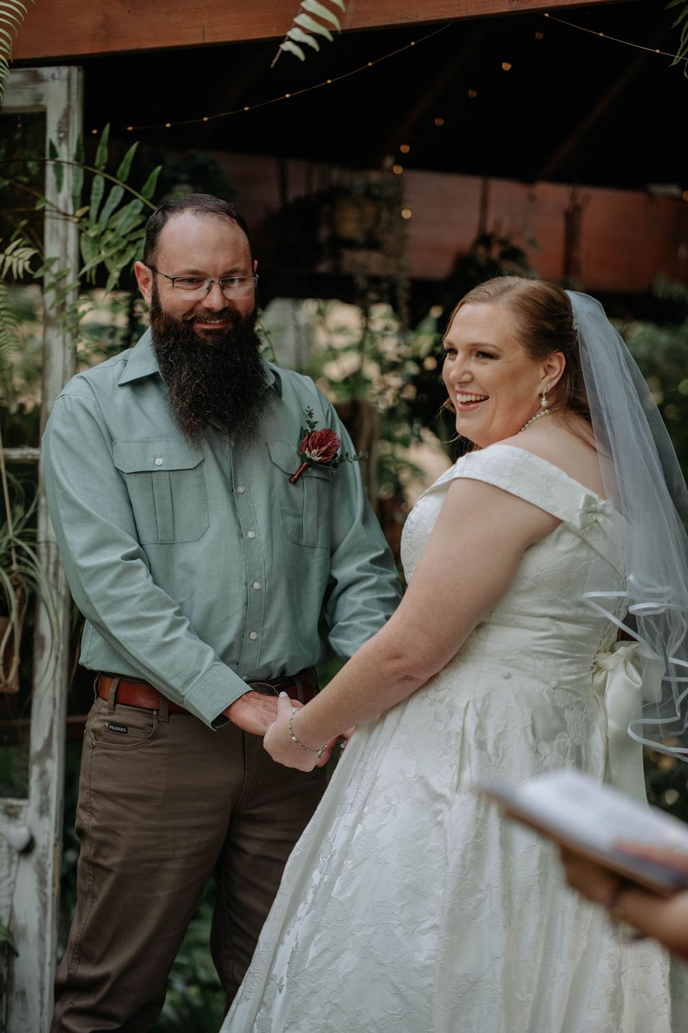 Bride in white off-shoulder gown and veil laughs with bearded groom in sage shirt during outdoor wedding ceremony.