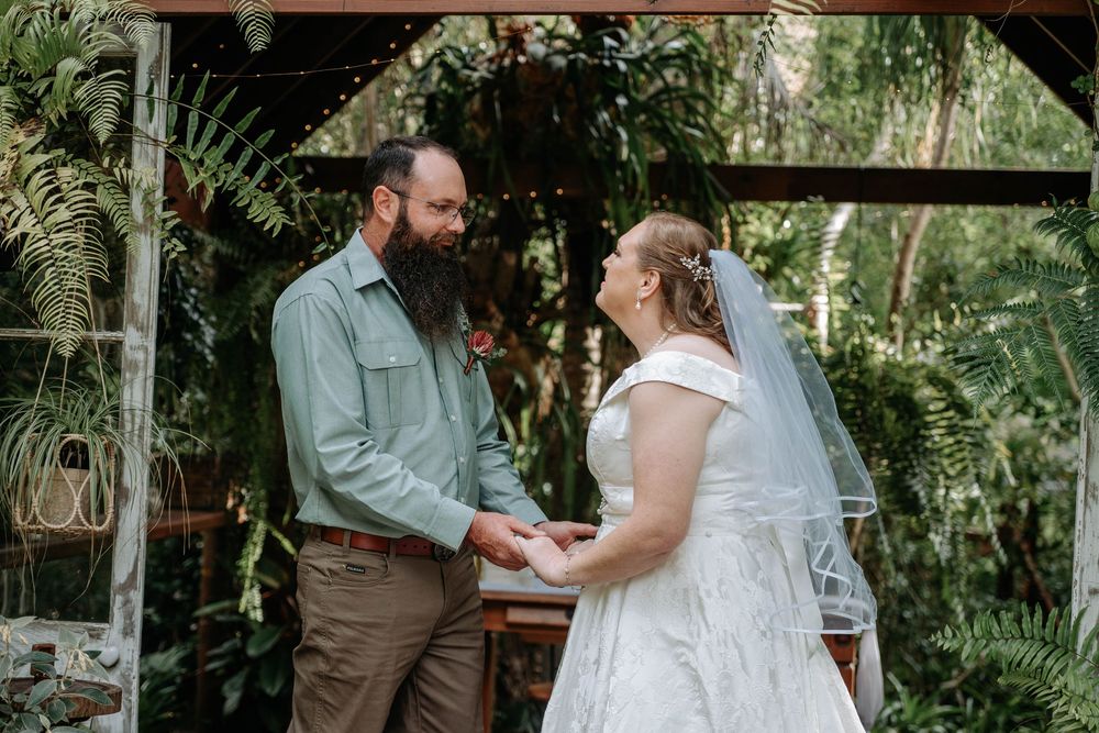 A bearded groom in green shirt holds hands with a bride in white lace dress and veil at a lush garden wedding ceremony.