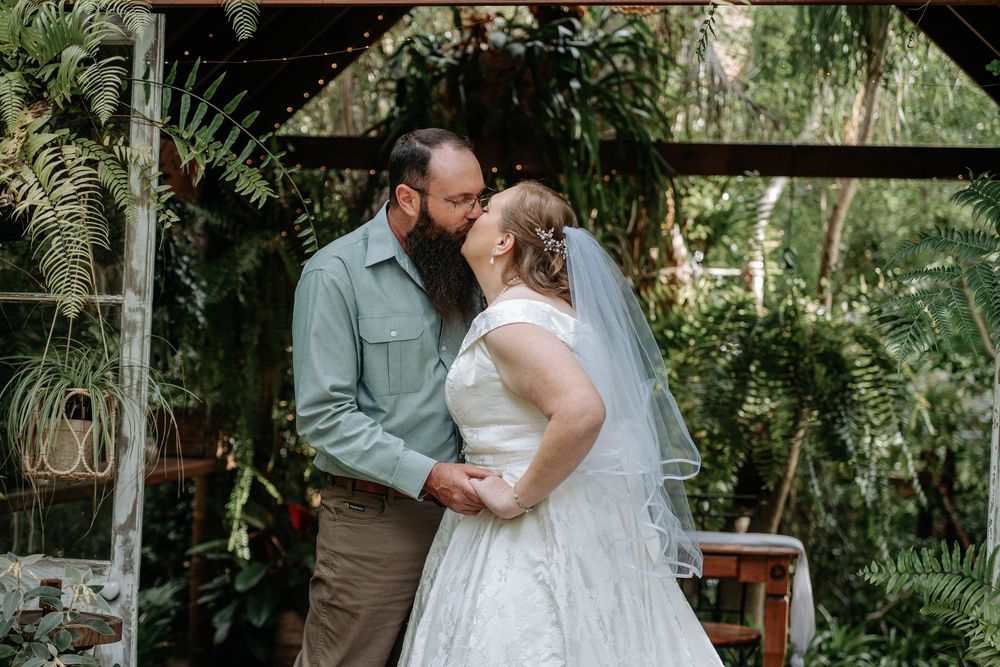 A couple shares a kiss at their outdoor wedding ceremony surrounded by lush tropical greenery, bride in white gown and veil.