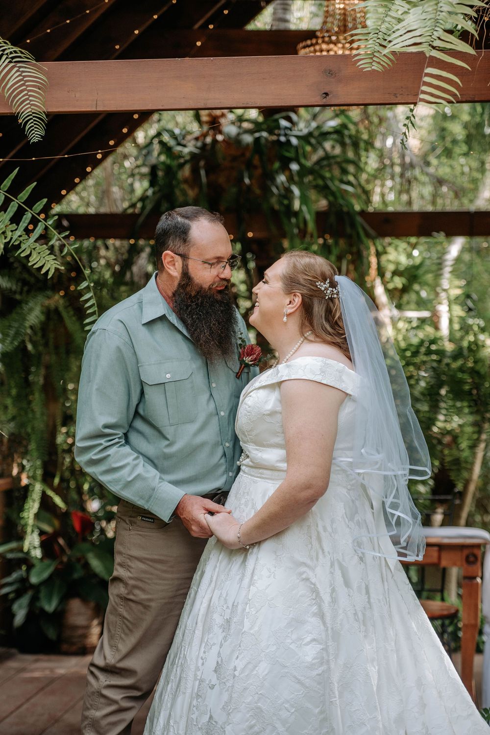 Bride in white lace gown and veil gazes lovingly at bearded groom in sage shirt during outdoor garden wedding ceremony.