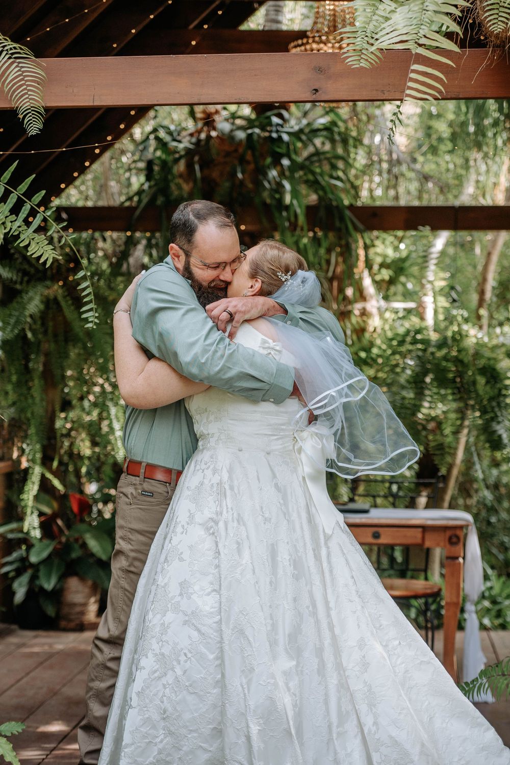 Bride and groom share a heartfelt embrace under a wooden pergola surrounded by lush greenery at an outdoor wedding.