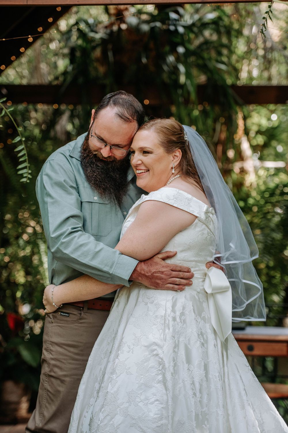 A bearded groom in a sage shirt embraces his smiling bride in a white gown and veil at an outdoor wedding ceremony.