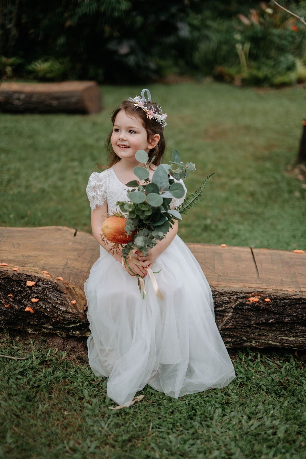 Young flower girl in white tulle dress holding eucalyptus bouquet, seated on rustic log in garden setting.