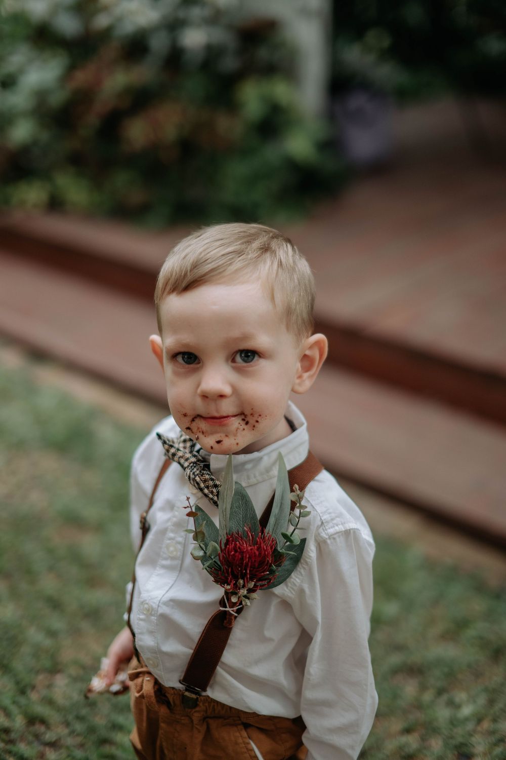 Adorable young ring bearer in white shirt and suspenders wearing a red floral boutonniere at an outdoor wedding.