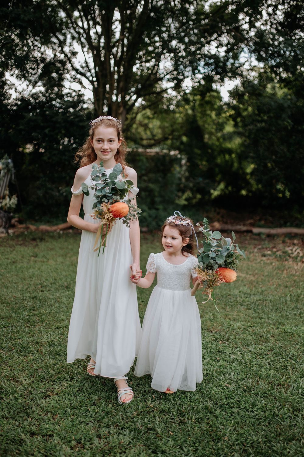 Two flower girls in white dresses holding orange floral bouquets with eucalyptus, posing outdoors on green grass.