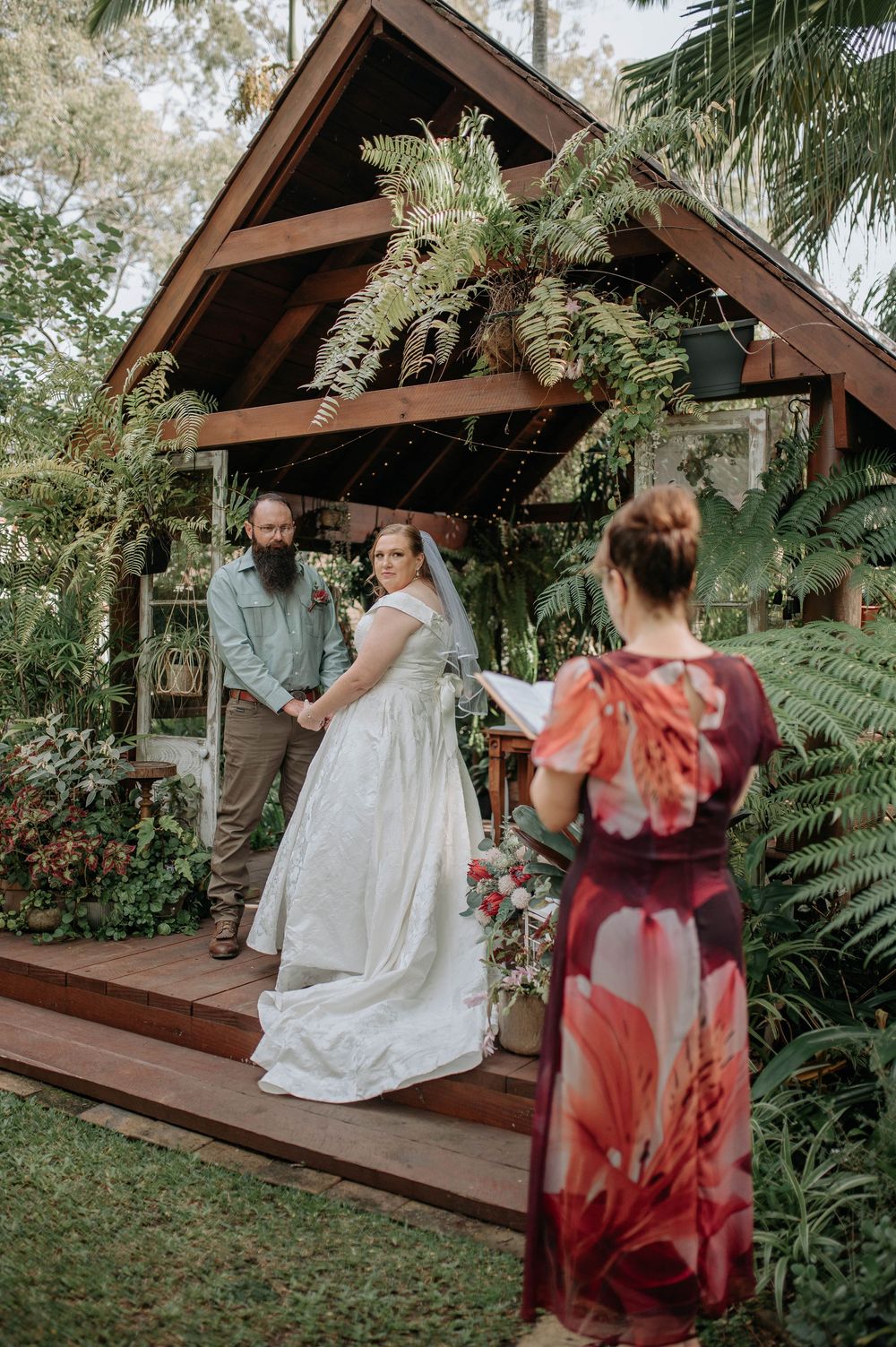 Bride and groom exchange vows at a lush garden wedding ceremony on a wooden deck surrounded by tropical ferns.