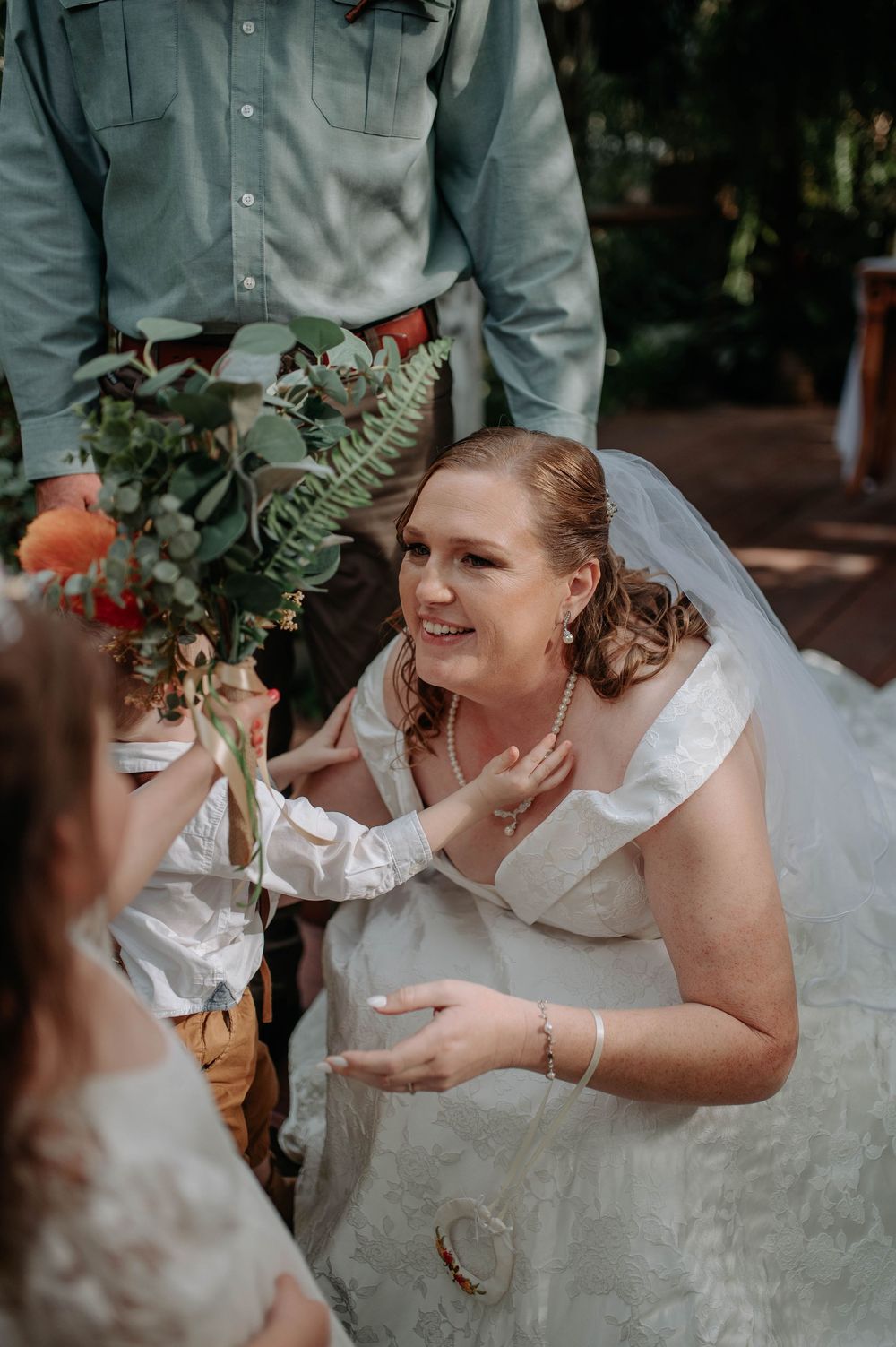 Bride in white gown smiles warmly at child holding bouquet during outdoor wedding ceremony.