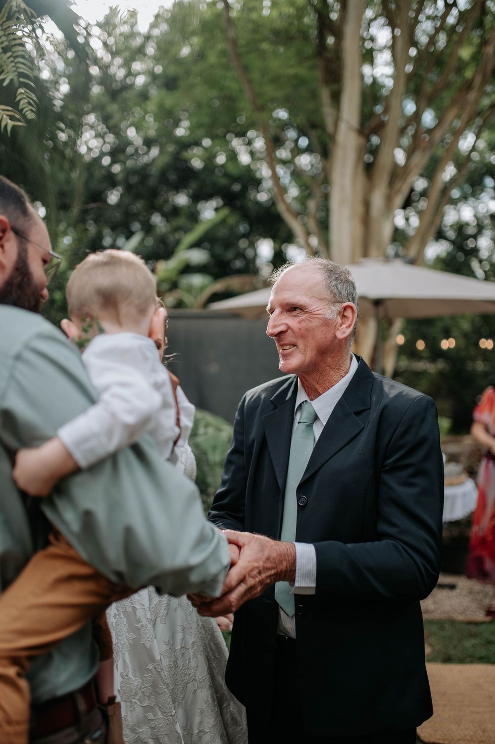 Elderly man in dark suit and mint tie shaking hands at an outdoor wedding reception with trees in background.