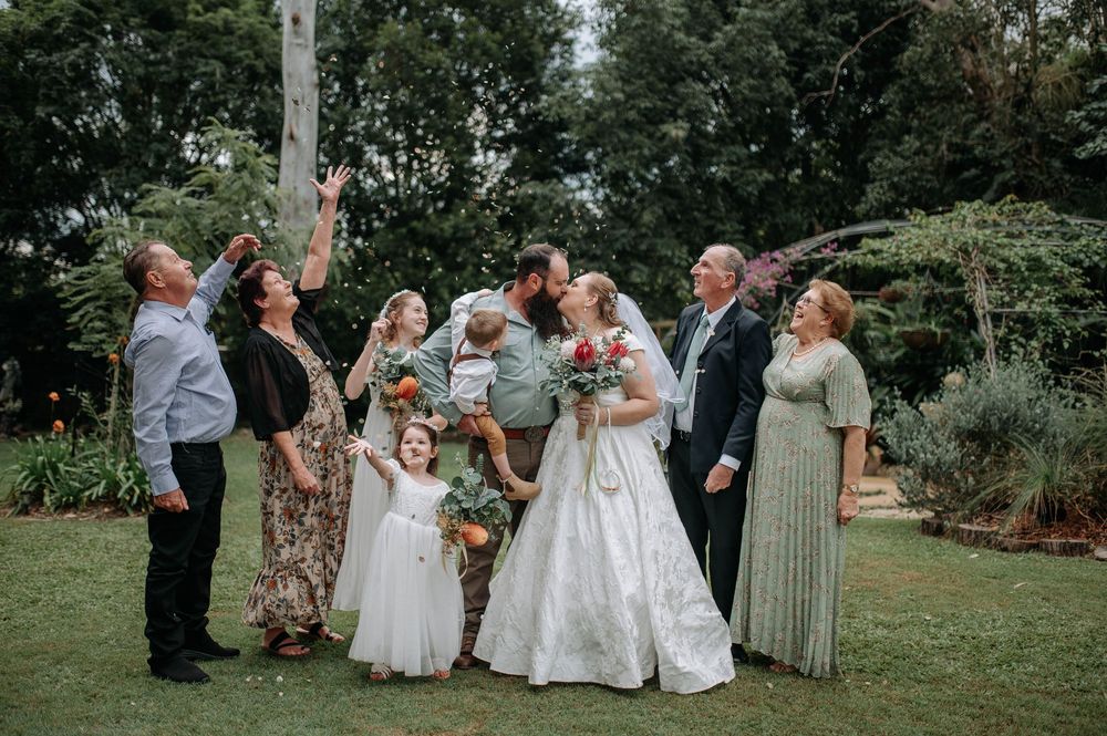 Wedding couple kissing outdoors surrounded by family in lush garden setting, bride in white gown, guests celebrating.