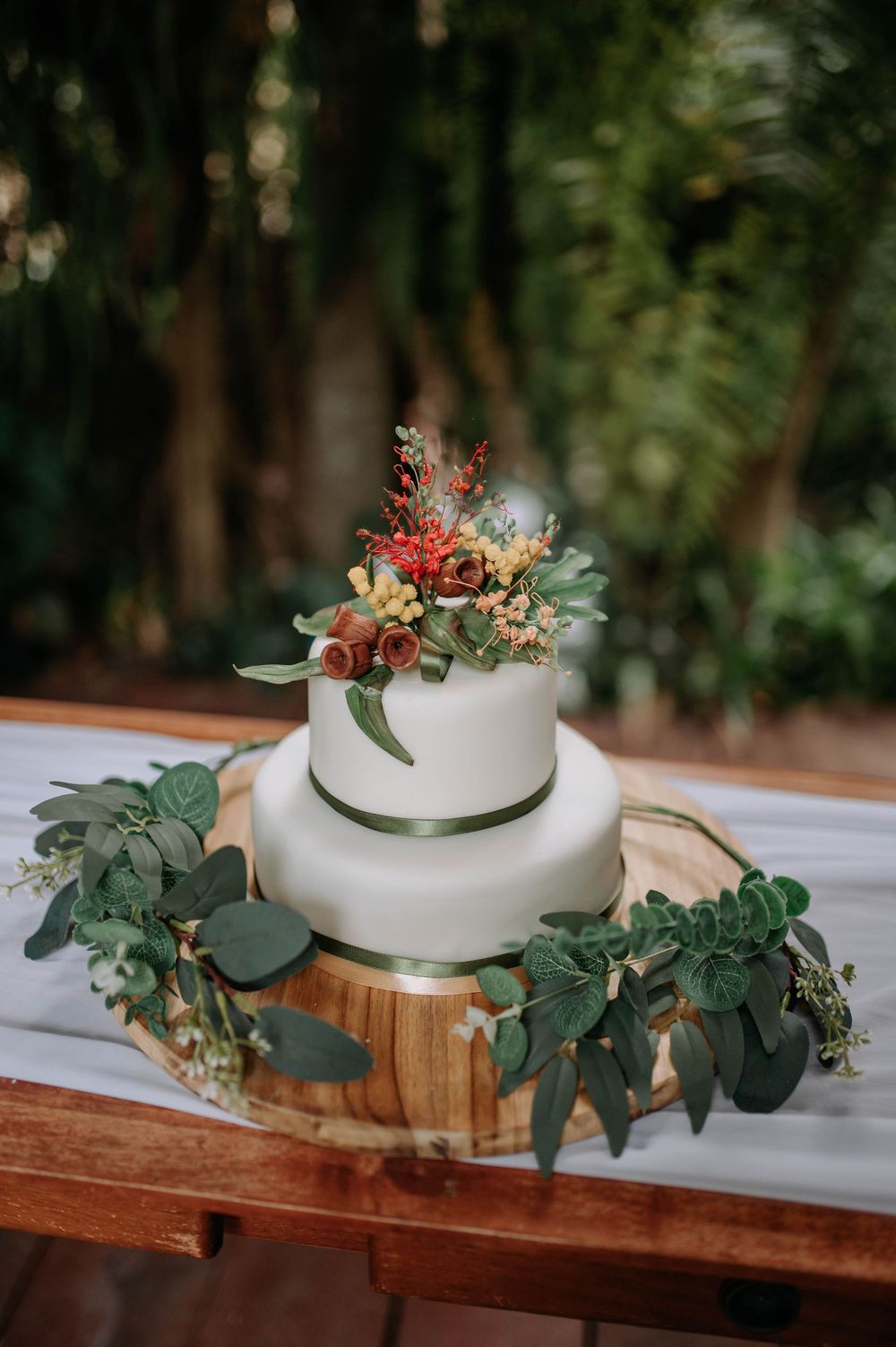 Two-tier white wedding cake with red and green floral topper on wood stand, surrounded by eucalyptus greenery.