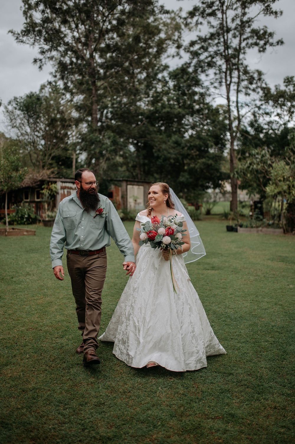Bride in white gown holding bouquet walks hand in hand with bearded groom in blue shirt on green lawn.