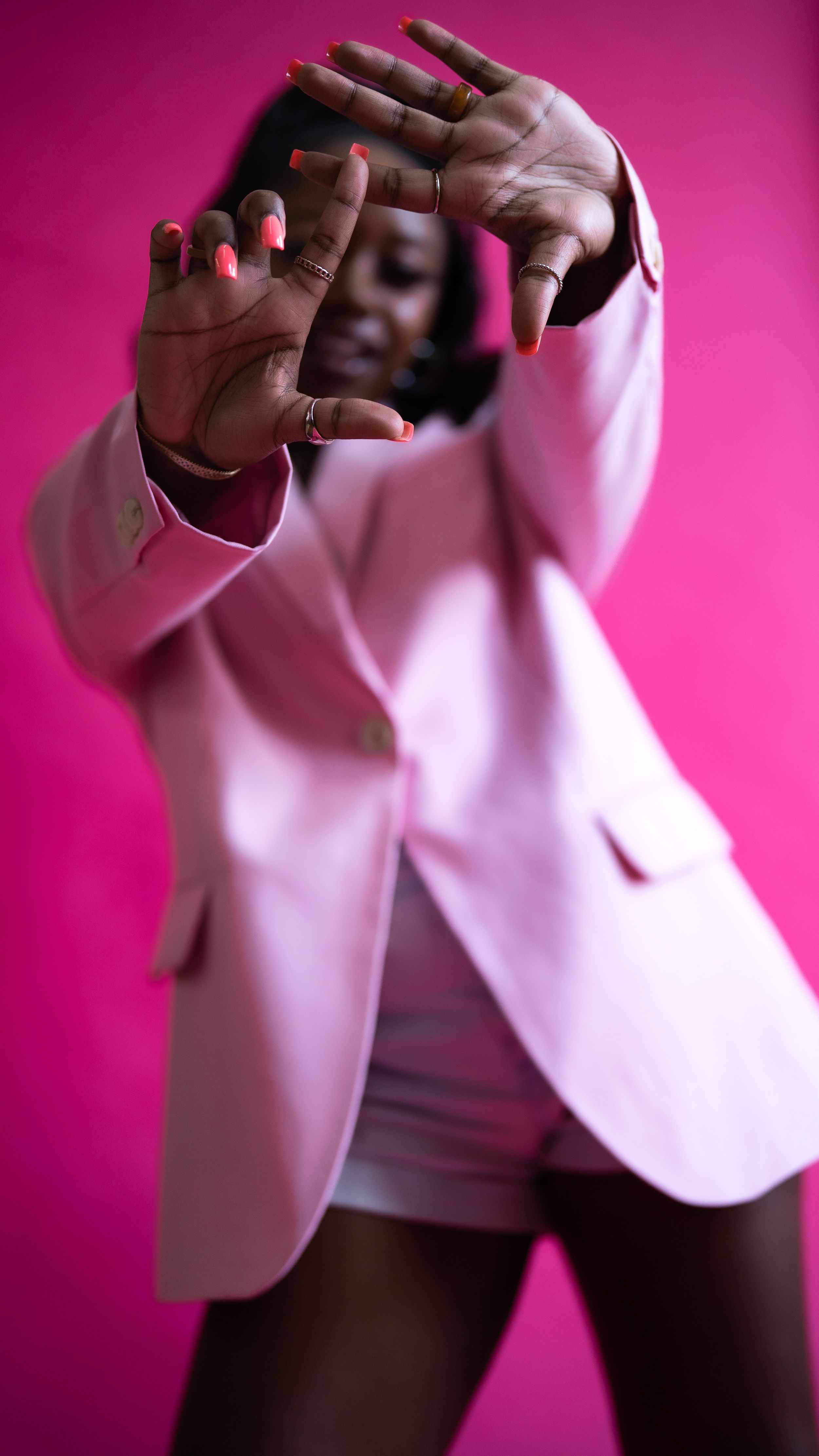 A Black woman in a pink blazer poses with hands outstretched toward camera against a vibrant pink background.