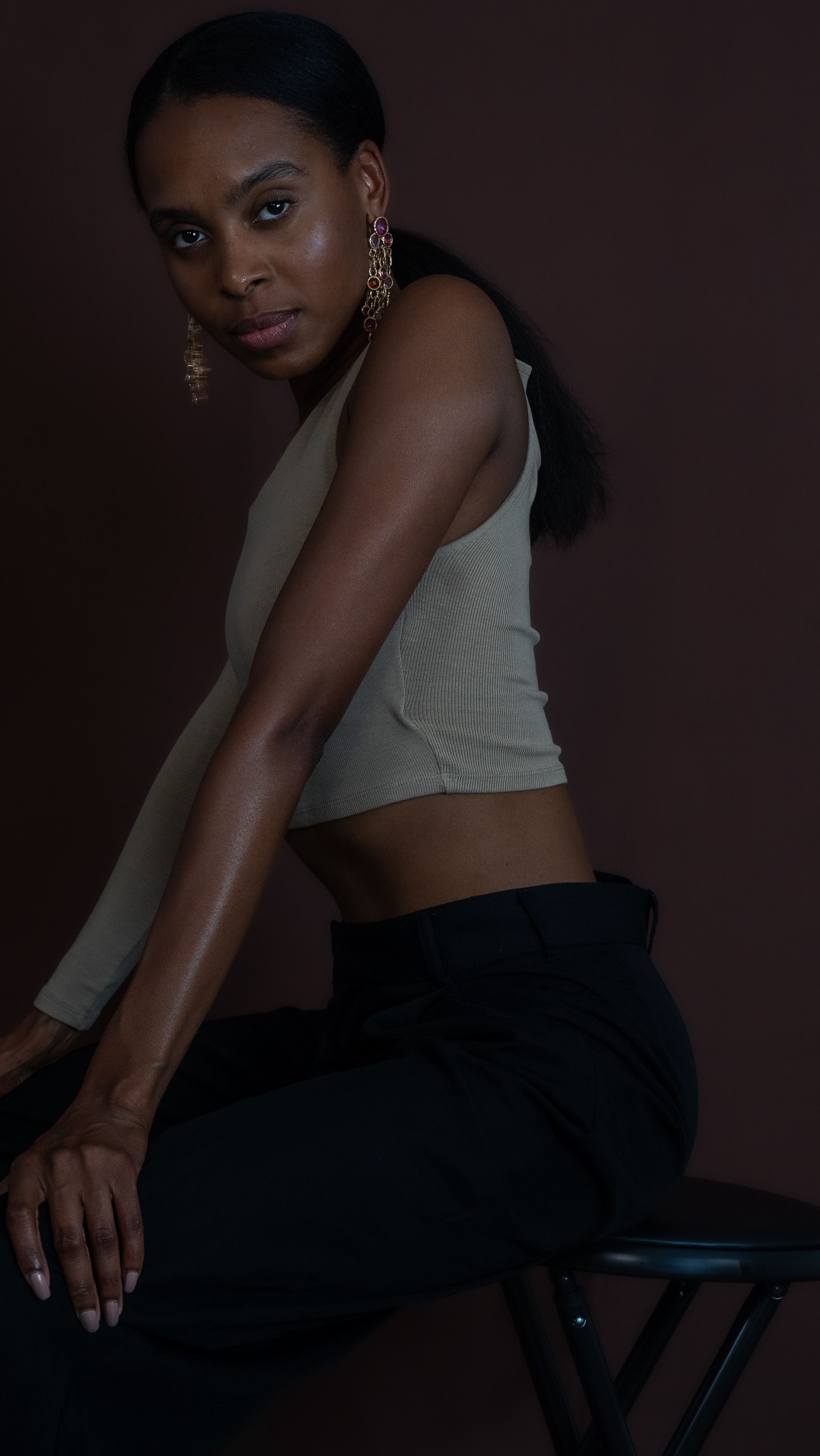 A Black woman in a beige crop top and black pants sits confidently on a stool against a dark background.