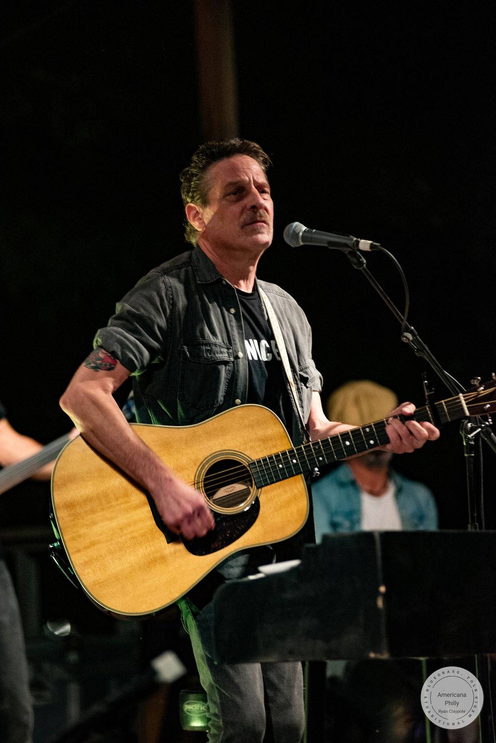 A male musician plays acoustic guitar and sings into a microphone on stage wearing a dark jacket.