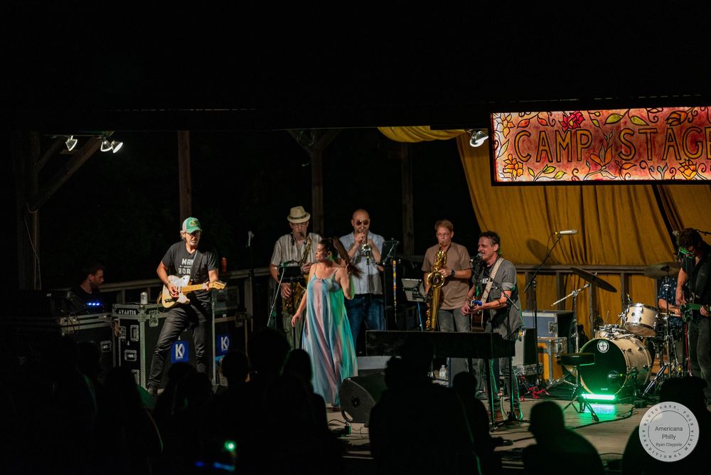 Live band performing on outdoor stage at night under colorful Camp Stage banner with multiple musicians.