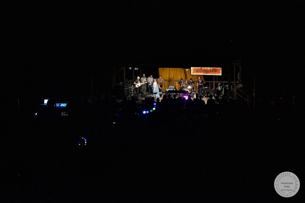Dark concert venue with performers on a lit stage surrounded by audience silhouettes at an outdoor night show.