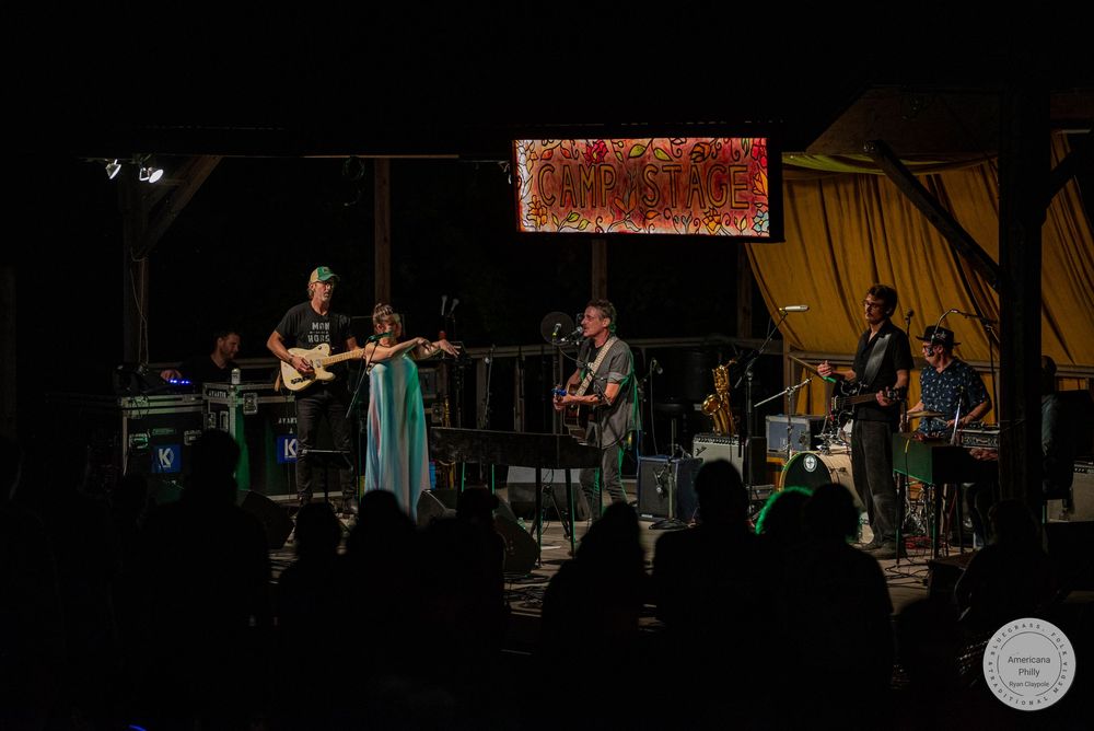 Live band performing on a dimly lit outdoor stage with colorful backdrop lighting at a nighttime concert event.