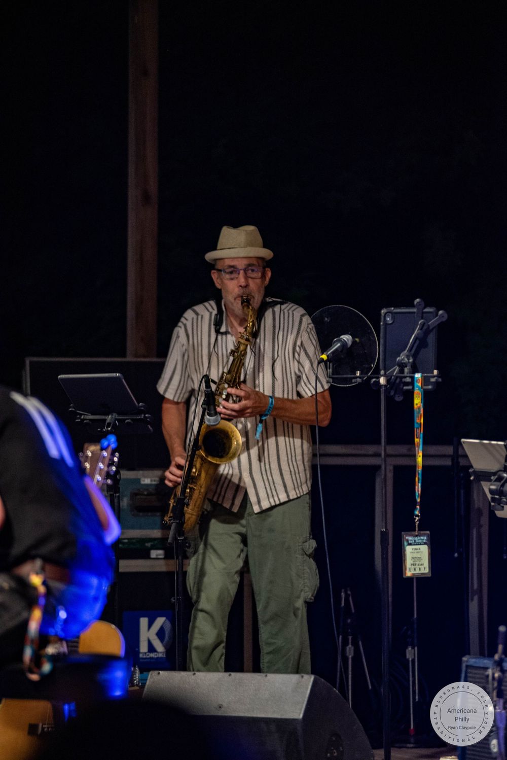 Man in striped shirt and fedora playing saxophone on outdoor stage at night music festival.