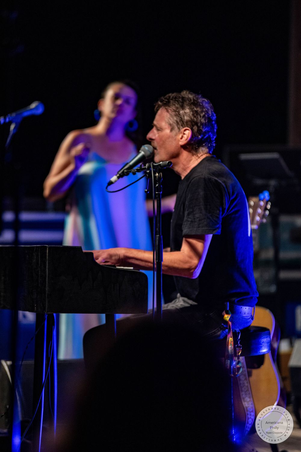 A man plays keyboard and sings on stage while a woman in a blue dress performs behind him.
