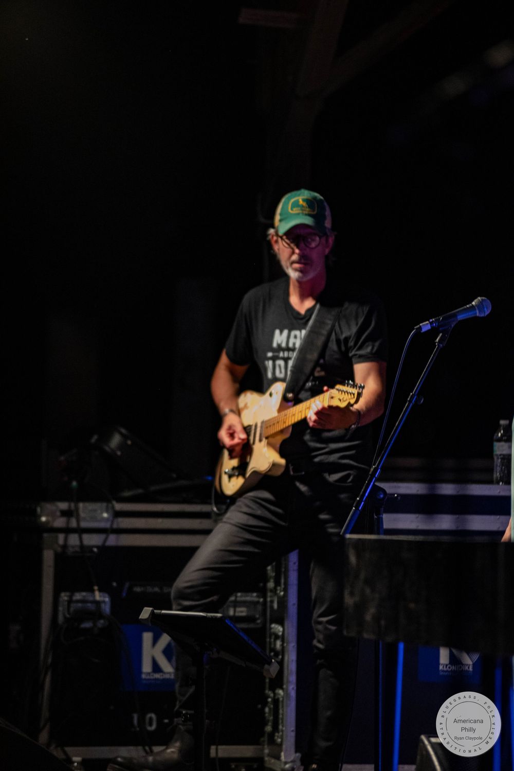 Guitarist performing on stage wearing a green cap and black t-shirt, playing an electric guitar under concert lighting.