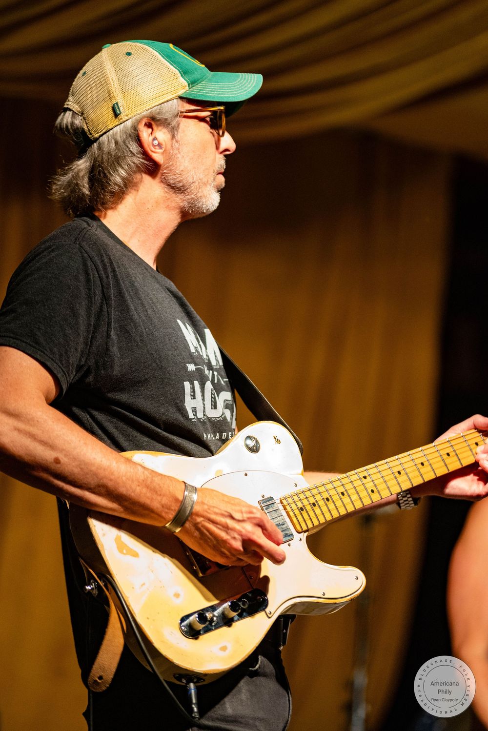Man playing white electric guitar on stage, wearing green cap and sunglasses, performing live music outdoors.