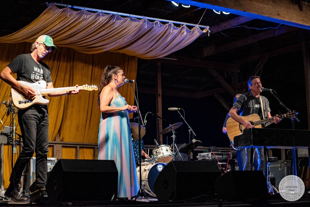 Live band performing on stage with guitarist, female vocalist in blue dress, and acoustic guitarist at outdoor venue.