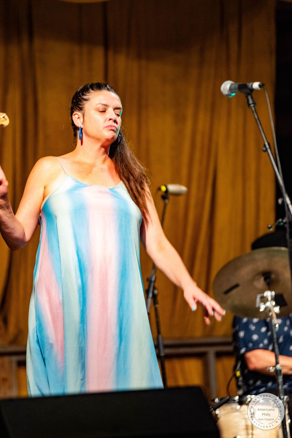 A woman in a colorful blue and pink dress performs on stage holding a maraca with eyes closed.