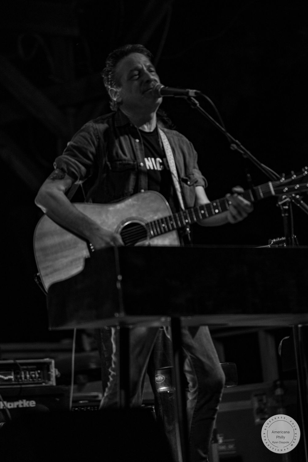 Black and white photo of a male musician playing acoustic guitar and singing into a microphone on stage.