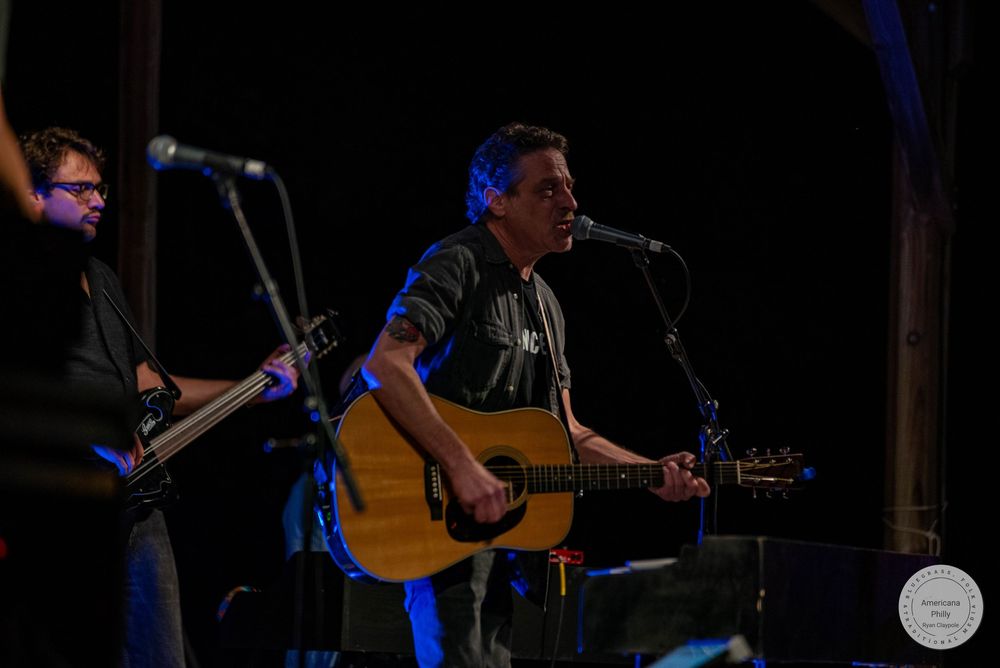 Musician playing acoustic guitar and singing into microphone on stage with guitarist performing in background.