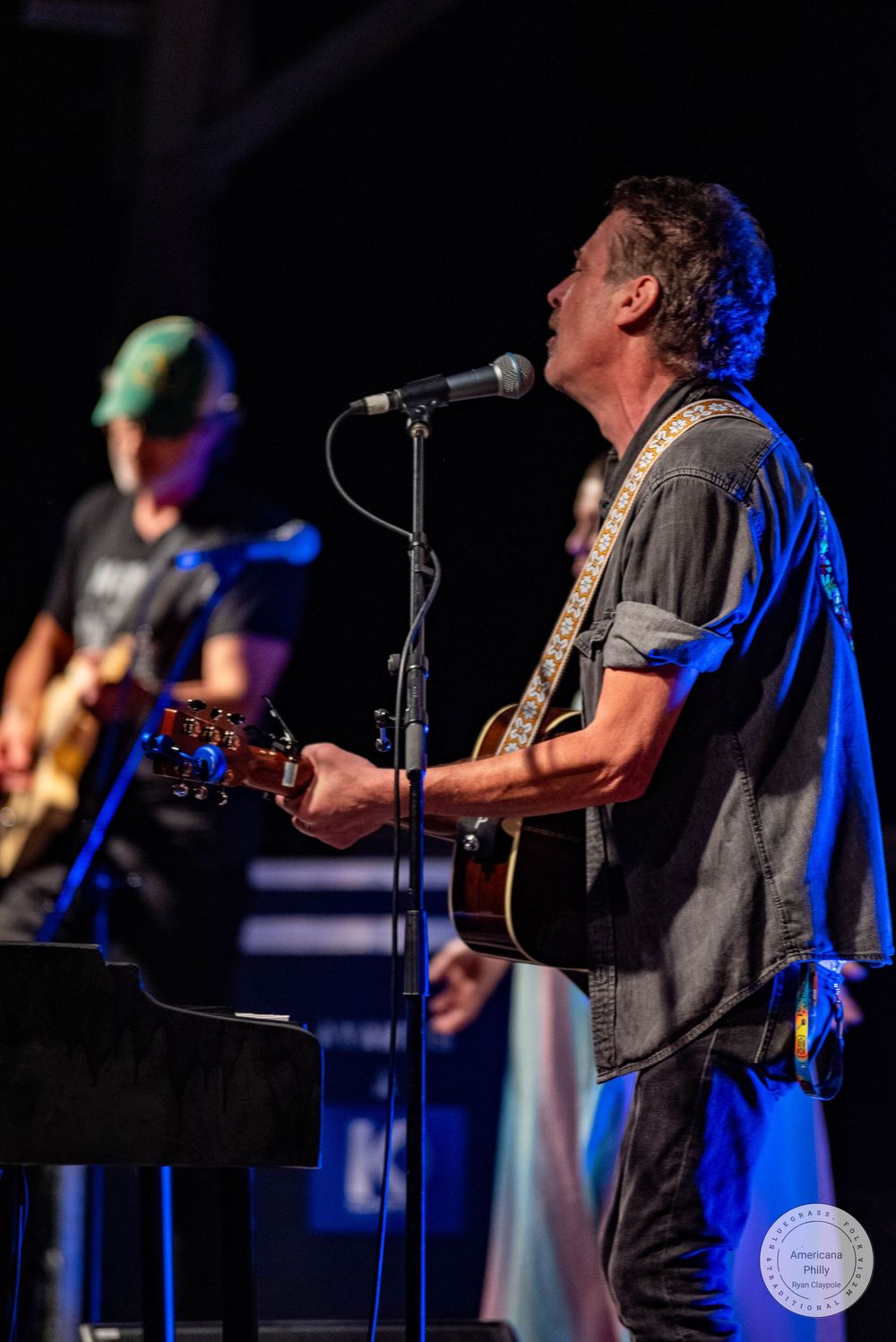 Musician singing into microphone while playing acoustic guitar on stage at outdoor concert.