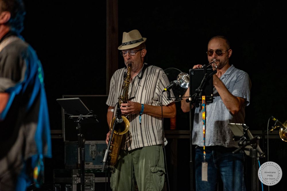 Two musicians perform live on stage, one playing saxophone and another playing trumpet at an outdoor concert.