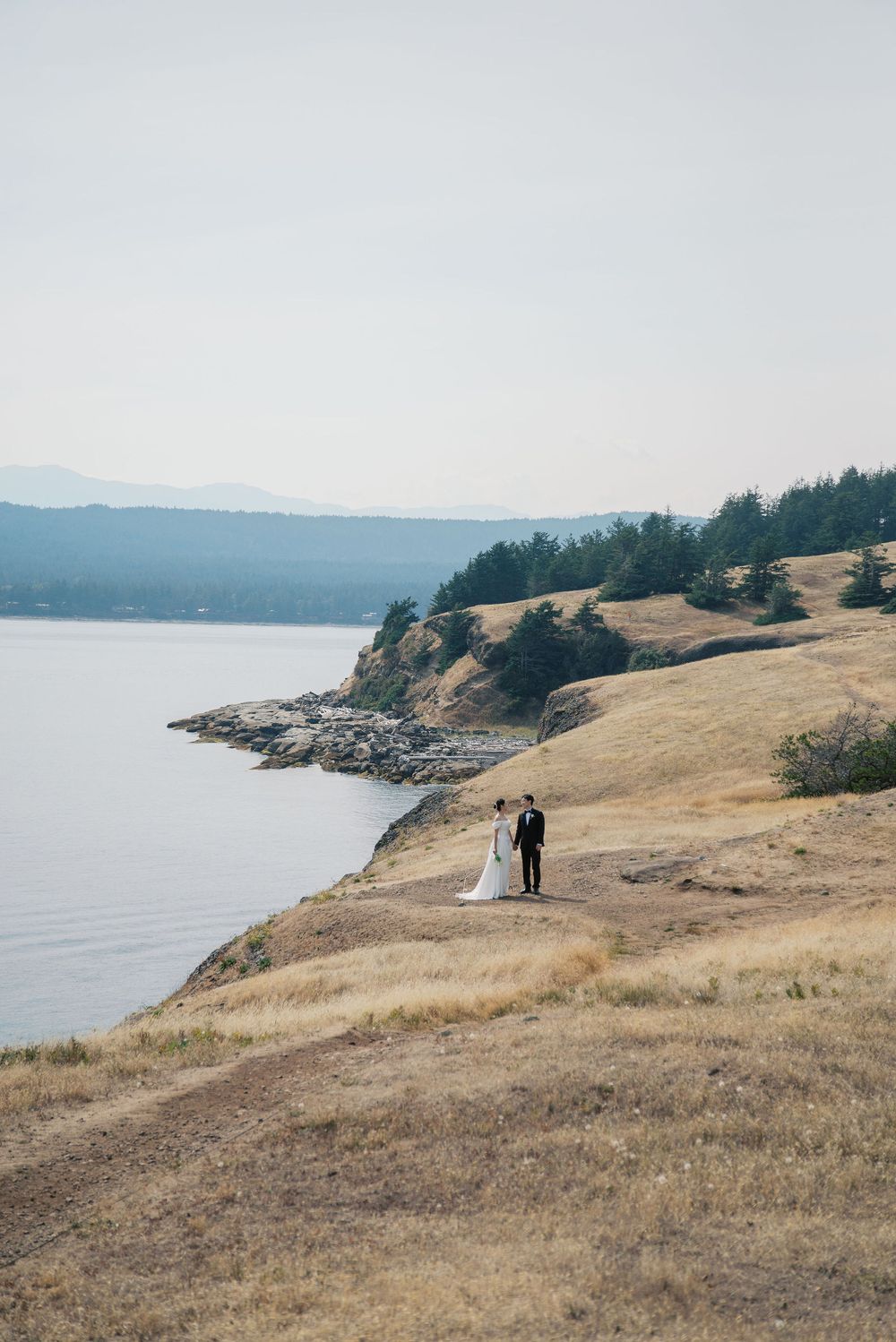 Bride and groom stand on golden coastal cliffs overlooking calm blue waters during their outdoor wedding photo session.