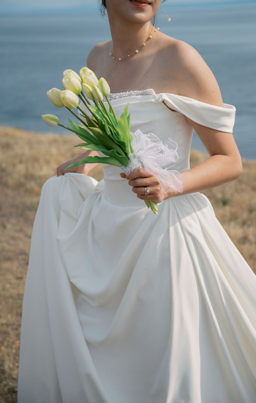 Bride in off-shoulder white gown holds bouquet of white tulips overlooking scenic coastal waters.