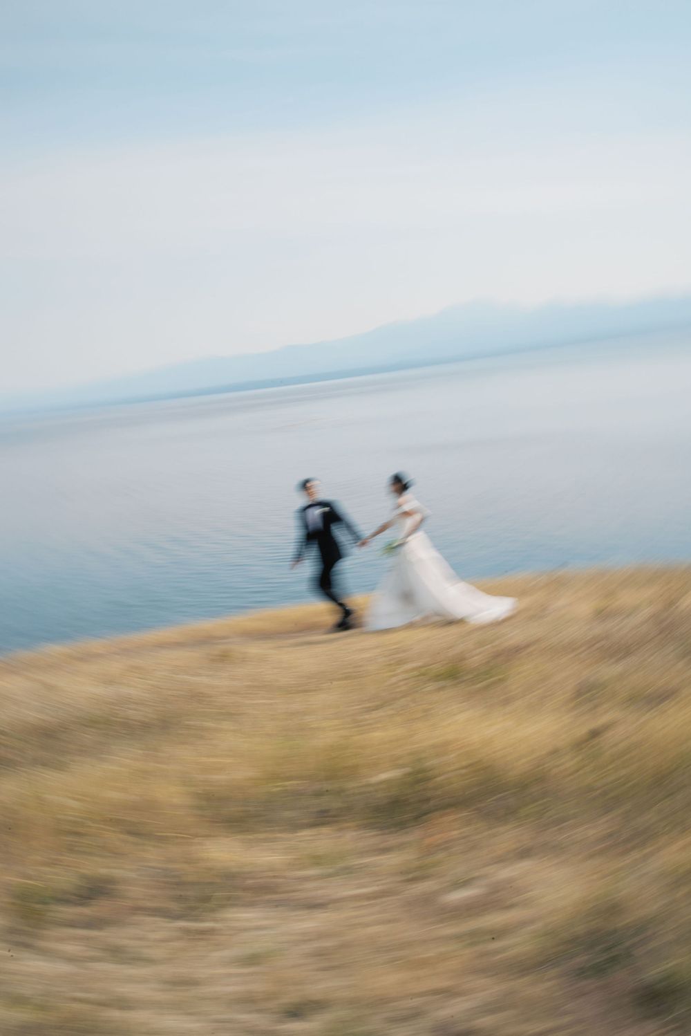 Blurry artistic wedding photo of bride and groom holding hands running along coastal cliffside with calm sea backdrop.