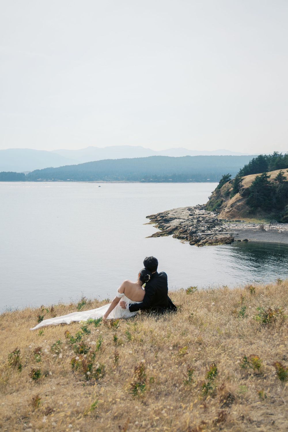 Wedding couple sit closely together on dry grass overlooking calm bay with forested rocky coastline.