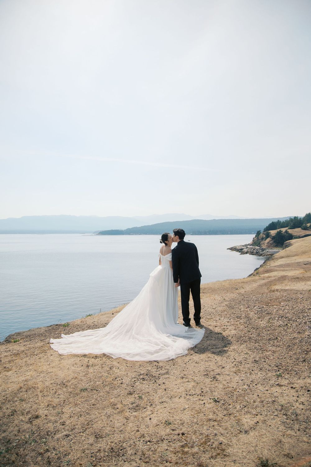 Bride and groom kissing in wedding attire on a rocky shoreline with calm blue water and misty mountains behind them.