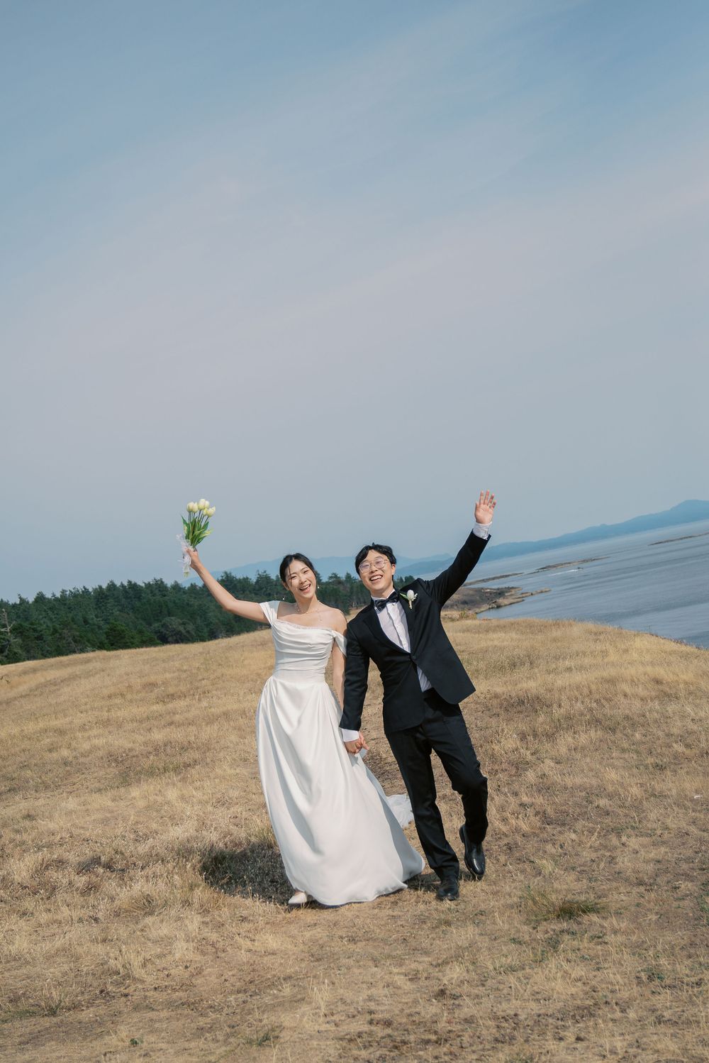Happy newlywed Asian couple celebrating on a scenic hillside, bride in white gown holding bouquet, groom in black tuxedo.