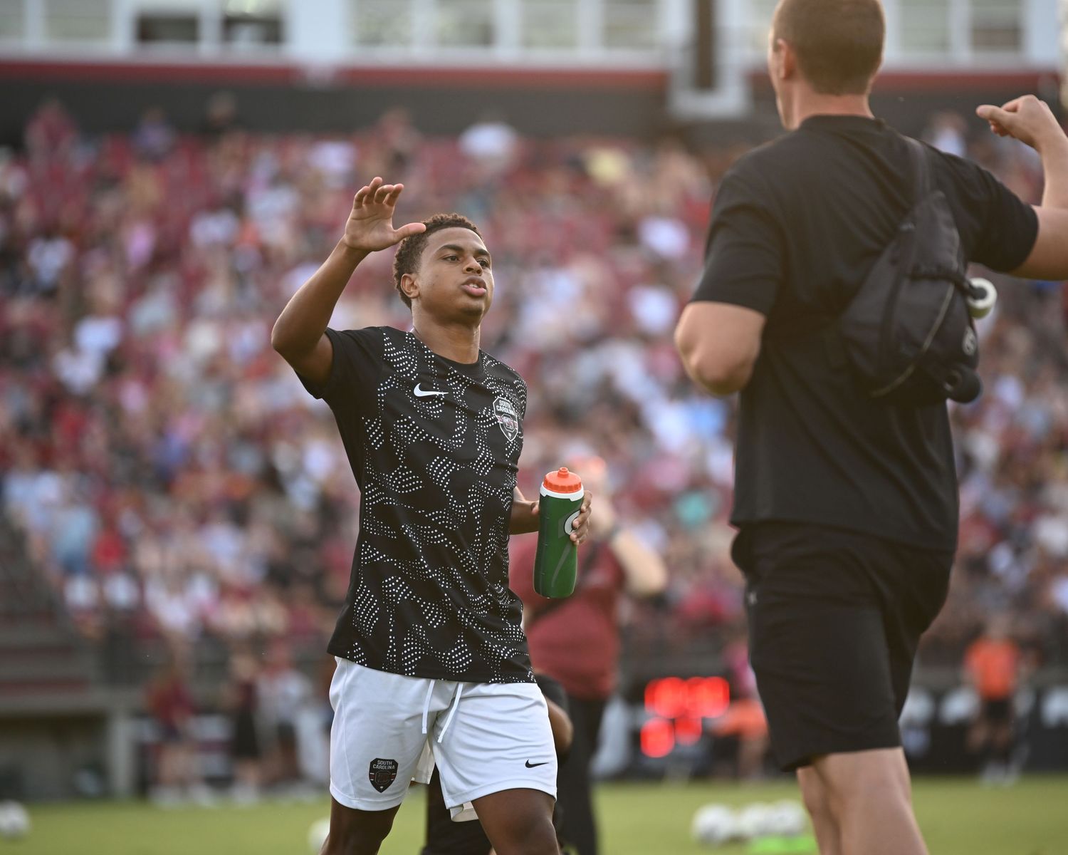 Soccer player in a black warmup shirt gestures toward a sideline camera.