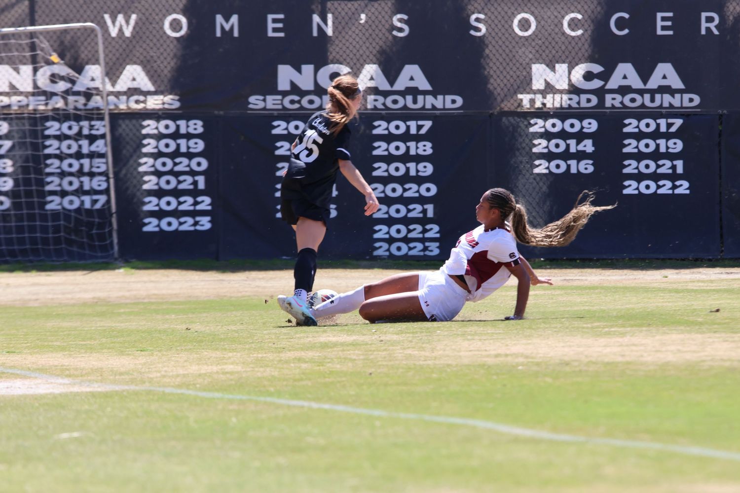 Women's soccer player slides toward the ball in front of NCAA tournament banners.