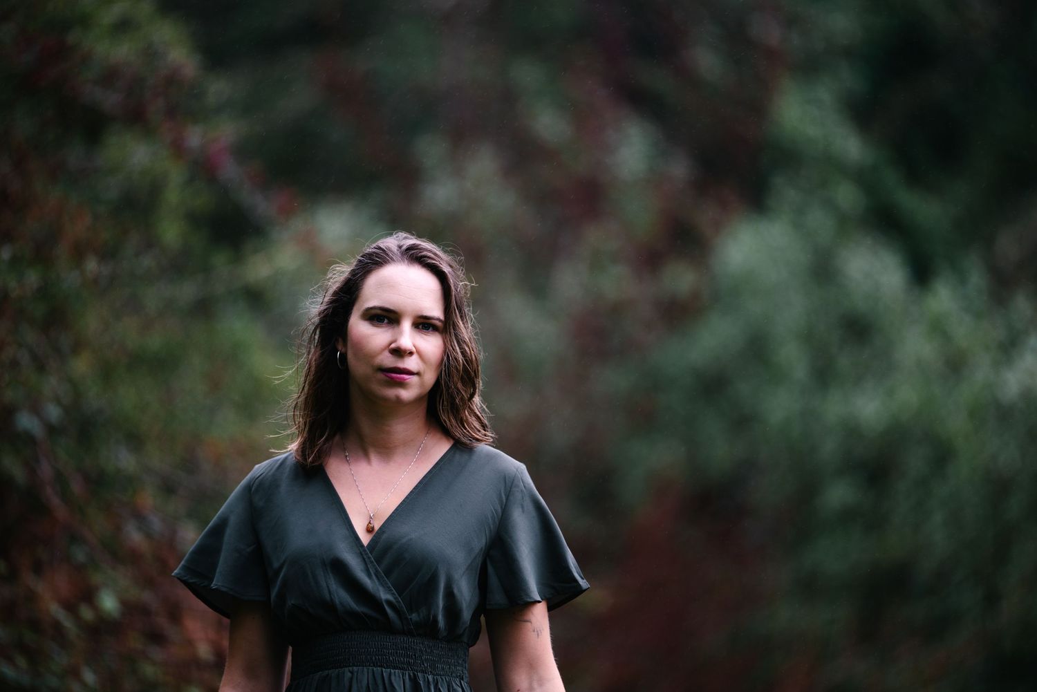 Woman in dark wrap dress standing outdoors among lush green and red foliage in a serene natural setting.