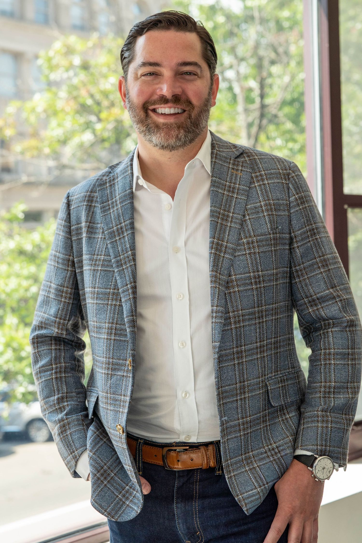 Smiling man with beard wearing plaid blazer and white shirt, standing near window with greenery background.
